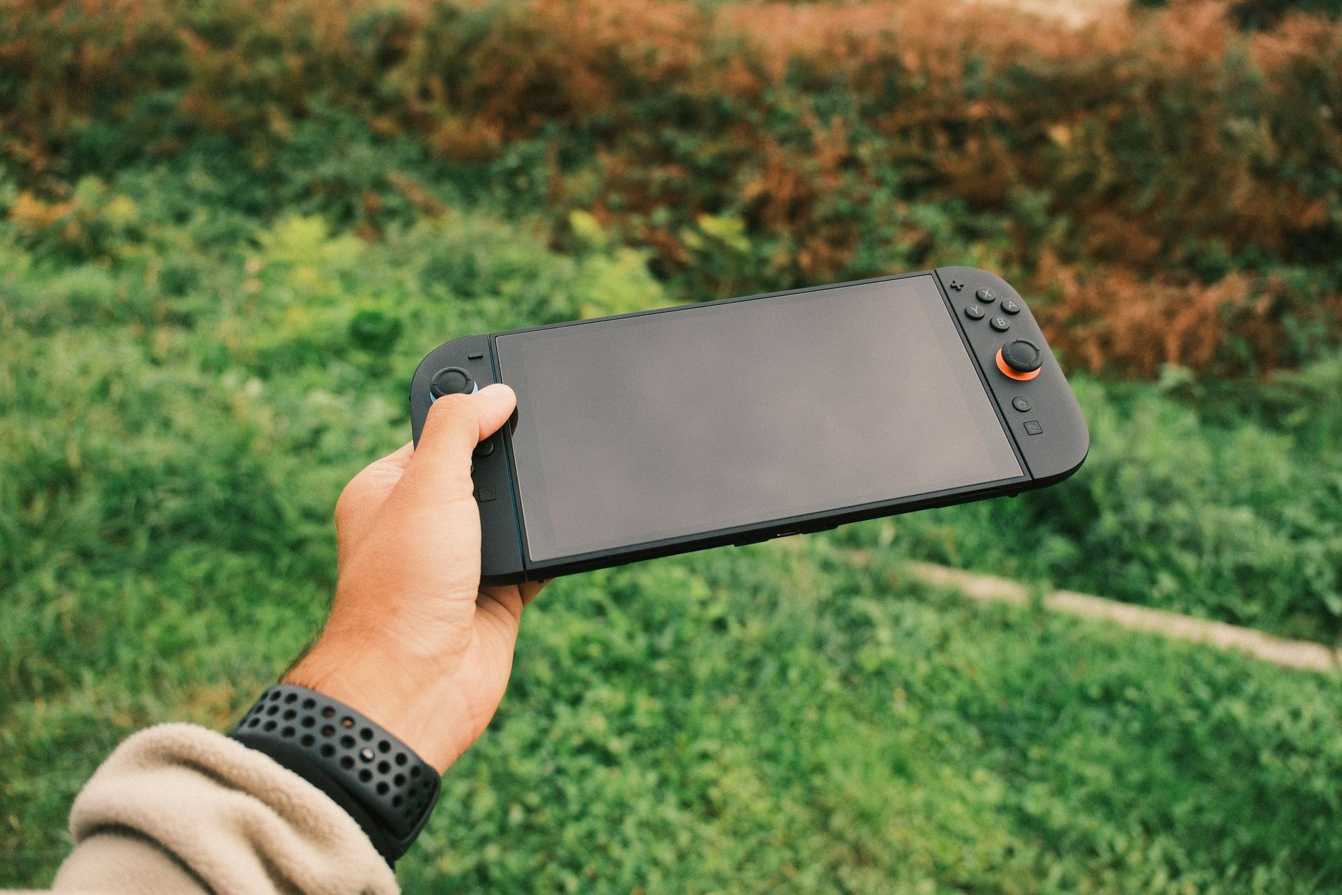Close-up of a person's hand holding a black handheld gaming console against a green outdoor background.